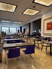 A spacious and modern dining area with neatly arranged tables and chairs. The room features a high ceiling with large, decorative light panels. The floor is polished with a reflective surface, and there's a large piece of artwork on the wall. Some people are visible in the background near the buffet area.