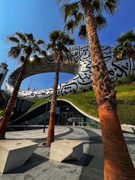 A futuristic building with large Arabic calligraphy on its exterior is framed by tall palm trees in the foreground. The structure features a unique torus shape, and its facade includes metallic elements and large glass windows. The surrounding area includes manicured green landscapes and modern geometric concrete benches on a paved walkway.