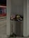 A metal trash bin labeled 'Abfall' is overflowing with various waste items such as paper cups, bottles, and bags. The surroundings appear to be a train or metro station, identifiable by the red train visible in the background. On the ground, near the base of the bin, are scattered flowers, including long-stemmed roses.