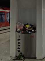 A metal trash bin labeled 'Abfall' is overflowing with various waste items such as paper cups, bottles, and bags. The surroundings appear to be a train or metro station, identifiable by the red train visible in the background. On the ground, near the base of the bin, are scattered flowers, including long-stemmed roses.
