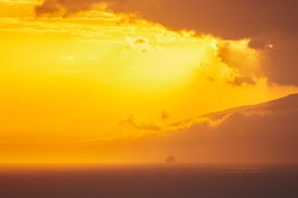 A vast expanse of ocean beneath a dramatic sunset sky. The sun casts a golden hue across the scene, with scattered clouds and distant mountains creating depth. A cruise ship is silhouetted against the horizon, adding a sense of scale and tranquility.