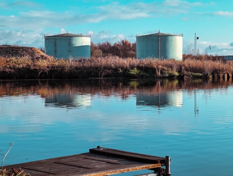 Two large cylindrical storage tanks are located near a body of water with a clear reflection. The tanks are surrounded by dry grass and vegetation, and the scene includes a partly cloudy sky with blue tones.