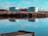 Two large cylindrical storage tanks are located near a body of water with a clear reflection. The tanks are surrounded by dry grass and vegetation, and the scene includes a partly cloudy sky with blue tones.