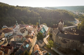 An aerial view of a historic town with quaint, colorful buildings and a prominent stone castle. The town is nestled among lush, green forests and surrounded by hills. A bridge extends over a river in the background. The architecture features old-world charm with steep, gabled roofs and a central cobblestone pathway.