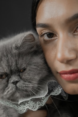A close-up photograph featuring a person and a gray Persian cat. The cat has a fluffy fur coat and is wearing a lace accessory. The person is partially visible, with an emphasis on the eyes and lips. Both appear to be in a relaxed pose.