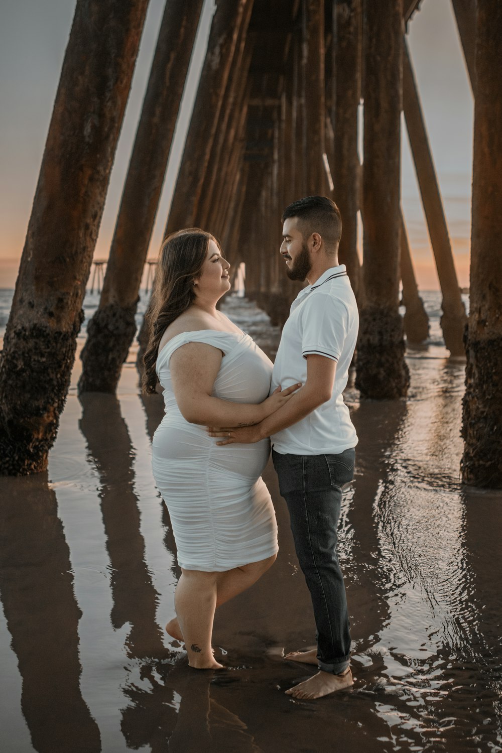 a pregnant couple standing under a pier at sunset