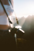 A person stands with a camera in focus while the background reveals a scenic view of mountains bathed in soft sunlight. The image is slightly blurred, creating a dreamy and natural ambiance.