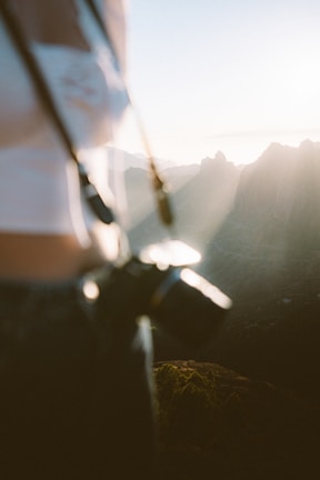 A person stands with a camera in focus while the background reveals a scenic view of mountains bathed in soft sunlight. The image is slightly blurred, creating a dreamy and natural ambiance.