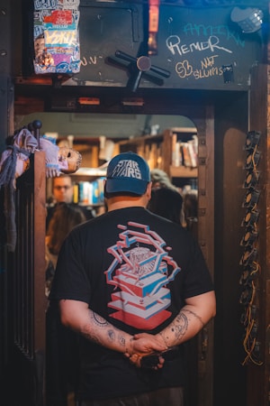 A person wearing a 'Star Wars' cap stands in a doorway with their back to the viewer. The person's t-shirt features a colorful, geometric design. The doorway is adorned with graffiti and stickers, including a doll attached to the side. Inside, bookshelves are visible, creating a cozy, cluttered atmosphere.