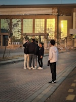 A group of people standing on a brick-paved walkway in front of a modern building with reflective glass windows. The individuals appear to be engaged in conversation, with one person slightly apart, observing the group. The setting is at sunset, casting a warm glow on the scene.