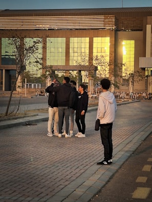 A group of people standing on a brick-paved walkway in front of a modern building with reflective glass windows. The individuals appear to be engaged in conversation, with one person slightly apart, observing the group. The setting is at sunset, casting a warm glow on the scene.