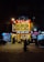 A brightly lit restaurant with a red sign attracts attention on a dimly lit street. The establishment is bustling with people gathered at the entrance and inside. Adjacent shops feature various signs, adding to the urban atmosphere.