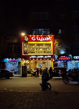 A brightly lit restaurant with a red sign attracts attention on a dimly lit street. The establishment is bustling with people gathered at the entrance and inside. Adjacent shops feature various signs, adding to the urban atmosphere.