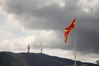 A large flag with red and yellow colors waves on a flagpole in the foreground. In the background, there is a mountainous landscape with a large cross structure and a telecommunications tower at the peak. The sky is overcast with dark, heavy clouds.