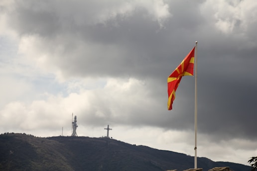 A large flag with red and yellow colors waves on a flagpole in the foreground. In the background, there is a mountainous landscape with a large cross structure and a telecommunications tower at the peak. The sky is overcast with dark, heavy clouds.
