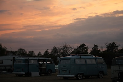 Several vintage vans are parked in an open area under a vibrant sunset sky. The vehicles are classic models, possibly from the 1970s, characterized by their boxy shape and distinctive design. In the background, a few trees and a trailer are visible, creating a serene and nostalgic atmosphere.