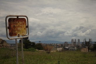 A weathered, rusted sign with inscriptions including 'DO NOT CROSS' stands in an open grassy field. In the background, a cityscape with several tall buildings is visible under a cloudy sky.
