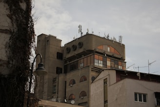 The image features a multi-story building with a unique architectural design. The structure includes several satellite dishes and antennas on the roof, indicating its use for telecommunications or broadcasting. The facade of the building displays circular and rectangular windows, some of which are tinted orange. Surrounding the building, there are additional structures and a street lamp, partially covered by ivy.