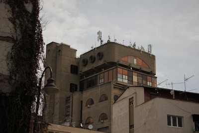 The image features a multi-story building with a unique architectural design. The structure includes several satellite dishes and antennas on the roof, indicating its use for telecommunications or broadcasting. The facade of the building displays circular and rectangular windows, some of which are tinted orange. Surrounding the building, there are additional structures and a street lamp, partially covered by ivy.