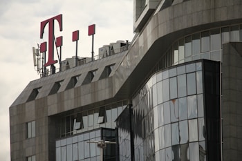 A modern office building with a glass facade features sharp, angular architectural lines. A large red 'T' logo is prominently displayed on the rooftop, accompanied by several antenna structures.