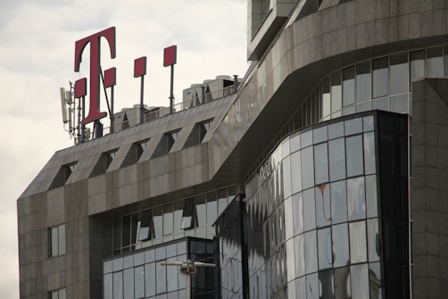 A modern office building with a glass facade features sharp, angular architectural lines. A large red 'T' logo is prominently displayed on the rooftop, accompanied by several antenna structures.