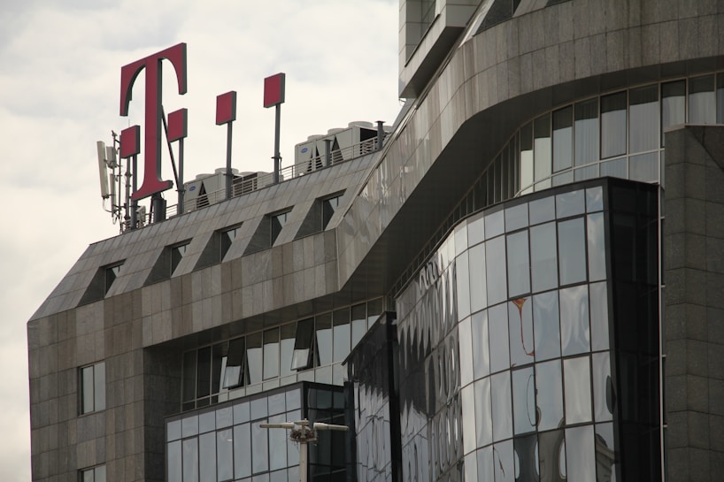 A modern office building with a glass facade features sharp, angular architectural lines. A large red 'T' logo is prominently displayed on the rooftop, accompanied by several antenna structures.