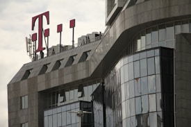 A modern office building with a glass facade features sharp, angular architectural lines. A large red 'T' logo is prominently displayed on the rooftop, accompanied by several antenna structures.