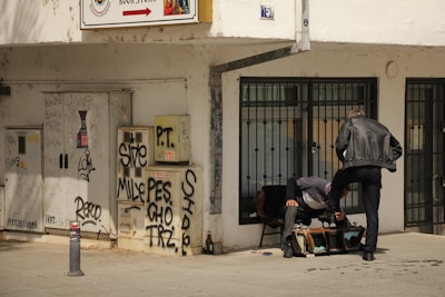 Two individuals are positioned outside a building with graffiti-covered walls and a storefront that has metal security bars. One person is seated, while the other leans forward with a foot on a chair, engaging in a conversation. The area is urban and appears somewhat neglected.
