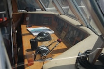 A boat cockpit with a wooden dashboard featuring various instruments and controls. There are several gauges and dials, and a throttle lever with a red handle. A nautical chart is spread out on the dashboard and a plastic cup is placed nearby. The area is enclosed by glass windows, through which light is streaming.