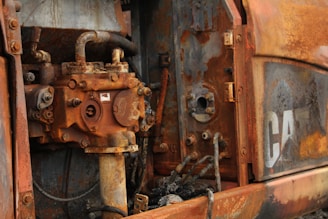 Close-up view of a heavily rusted and corroded industrial machine part, consisting of metal components with bolts, tubes, and pipes. The surface shows a range of rust colors, from deep orange to brown. Partially visible worn-off letters indicating a brand or logo.