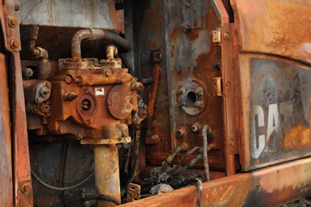 Close-up view of a heavily rusted and corroded industrial machine part, consisting of metal components with bolts, tubes, and pipes. The surface shows a range of rust colors, from deep orange to brown. Partially visible worn-off letters indicating a brand or logo.