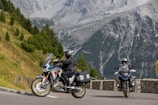 Motorcyclists are riding on a mountain road surrounded by lush greenery and steep rocky hillsides. The scene captures two motorcycles, with riders wearing protective gear, enjoying the picturesque landscape.