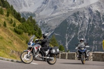 Motorcyclists are riding on a mountain road surrounded by lush greenery and steep rocky hillsides. The scene captures two motorcycles, with riders wearing protective gear, enjoying the picturesque landscape.