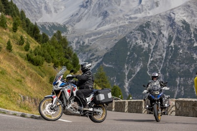 Motorcyclists are riding on a mountain road surrounded by lush greenery and steep rocky hillsides. The scene captures two motorcycles, with riders wearing protective gear, enjoying the picturesque landscape.