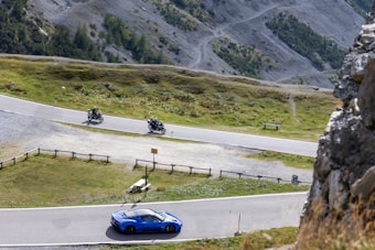 A bright blue sports car is driving along a winding mountain road. Two motorcyclists are riding on the adjacent road above, surrounded by a rocky and grassy landscape with sparse trees. The scene suggests a peaceful yet exhilarating journey through a mountainous area.