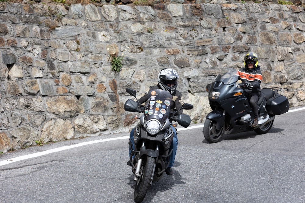 Dos motociclistas circulan por la carretera frente a un muro de piedra