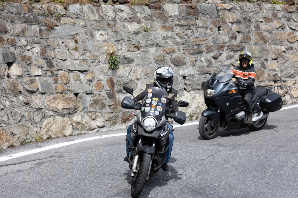 two motorcyclists are riding down the road in front of a stone wall