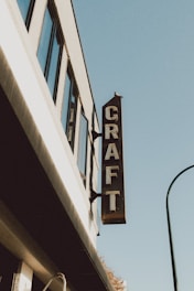 A building facade with a vertical sign reading 'CRAFT' protruding from it. The structure features several windows and is set against a clear blue sky.