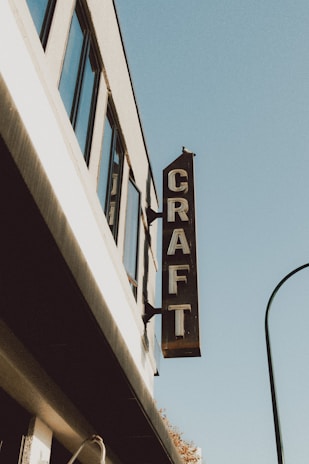A building facade with a vertical sign reading 'CRAFT' protruding from it. The structure features several windows and is set against a clear blue sky.