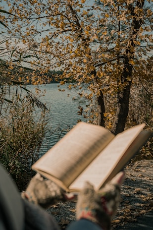 A smiling reader holding an open book in a sunlit park surrounded by autumn leaves.