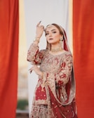 A woman dressed in an ornate red and gold traditional outfit adorned with intricate embroidery. She wears elaborate gold jewelry, including a choker, earrings, and a maang tikka. Her pose is graceful and elegant, with her hand raised near her face, and she has striking, well-applied makeup.