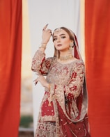A woman dressed in an ornate red and gold traditional outfit adorned with intricate embroidery. She wears elaborate gold jewelry, including a choker, earrings, and a maang tikka. Her pose is graceful and elegant, with her hand raised near her face, and she has striking, well-applied makeup.