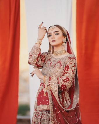 A woman dressed in an ornate red and gold traditional outfit adorned with intricate embroidery. She wears elaborate gold jewelry, including a choker, earrings, and a maang tikka. Her pose is graceful and elegant, with her hand raised near her face, and she has striking, well-applied makeup.