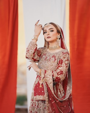 A woman dressed in an ornate red and gold traditional outfit adorned with intricate embroidery. She wears elaborate gold jewelry, including a choker, earrings, and a maang tikka. Her pose is graceful and elegant, with her hand raised near her face, and she has striking, well-applied makeup.