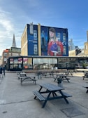 An urban area featuring a large outdoor seating space with multiple black picnic tables scattered on a concrete ground. In the background, a building displays a colorful mural with branding and a painted figure. Nearby, a tall spire and modern skyscrapers are visible under a partly cloudy sky.