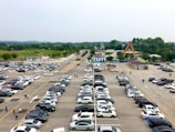 An expansive parking lot filled with a variety of cars, set against a backdrop of a park and amusement rides. The scene is lively with people walking amidst the rows of parked cars, and the park is lush with green trees.