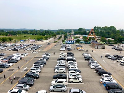 An expansive parking lot filled with a variety of cars, set against a backdrop of a park and amusement rides. The scene is lively with people walking amidst the rows of parked cars, and the park is lush with green trees.