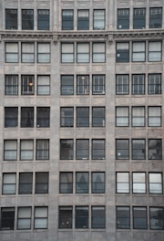 The image showcases a building facade with multiple windows arranged in a grid-like fashion. Each window has a dark frame and the building exterior is made of light-colored stone tiles. Some windows have shades drawn while others reveal partial views of interior lighting or objects.