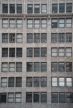 The image showcases a building facade with multiple windows arranged in a grid-like fashion. Each window has a dark frame and the building exterior is made of light-colored stone tiles. Some windows have shades drawn while others reveal partial views of interior lighting or objects.