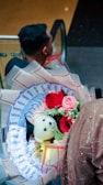 A bouquet featuring vibrant red and pink roses, a small teddy bear wearing a graduation cap, and decorative money arranged like flower petals is held by a person in a textured, light brown outfit. The background shows a person with a short hairstyle sitting in what appears to be an escalator setting.